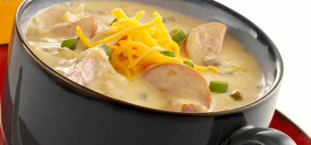A dark brown soup mug filled with Beer Cheese soup, garnished with chopped green onions and cheese shreds.