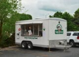 Food truck with a smiling vendor in an apron, serving ice cream, surrounded by greenery and parked in a lot.