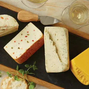 A cheese flight on a wooden and slate tray with four different varieties of cheese, a serving tool, and a glass of wine.