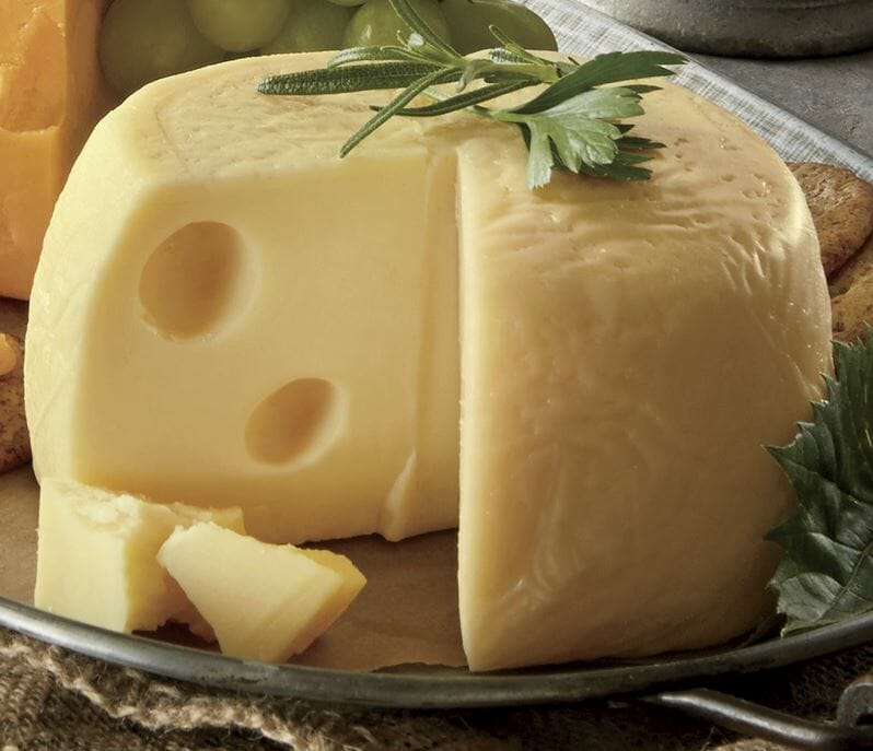 A round of buttery Baby Swiss cheese with a wedge removed to show the two distinctive holes, and herbal garnish on top.