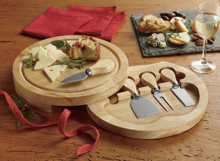 A wooden cheese board set with serving tools, an assortment of cheese, crackers, nuts, red napkins and a glass of white wine.