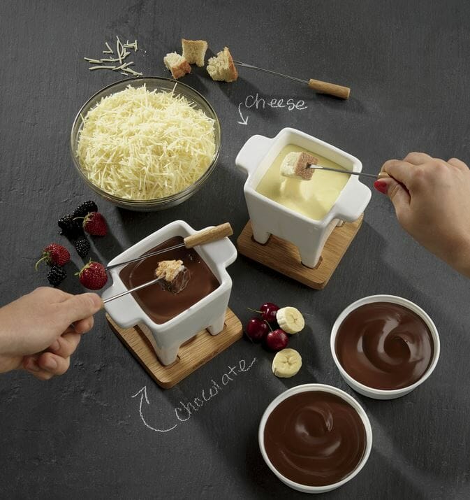 Two fondue pots of white cheese and chocolate for dipping bread chunks, berries, and banana slices with fondue forks.