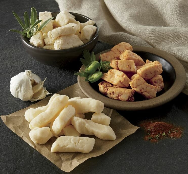 Three varieties of flavored cheese curds, two in bowls, one on brown paper, with a garlic bulb and herbs for garnish.