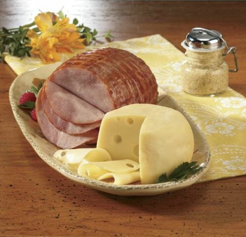 Spiral-sliced Ham with a round and slices of Baby Swiss Cheese on a platter with Dijon mustard and flowers on the side.
