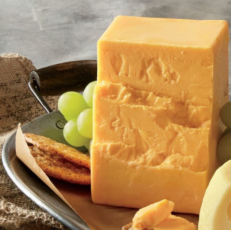 A rough sliced block of Sharp Cheddar cheese on a metal tray with green grapes and crackers.