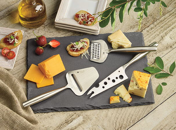 A slate cheese board displaying cheese tools with chunks and slices of cheese, strawberries, olive oil, and appetizers.