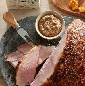 A seasoned ham with slices on a dark enameled platter, with a serving fork, tub of cranberry mustard, and cheese on the side.