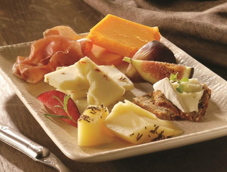 A square white plate displaying pairings of cheese varieties, figs, fruit, seeded bread, and pastrami thins.