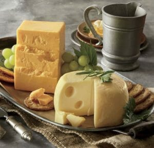 Sharp Cheddar cheese and a round of Baby Swiss cheese on a tray with green grapes and crackers, by a pewter mug.