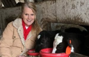 A lady farmer in a warm jacket sitting next to two Holstein calves feeding from two red buckets.