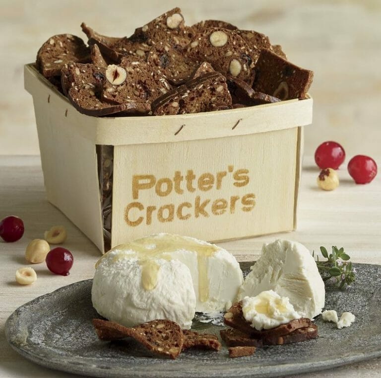 A basket of dark Potter's Crackers next to Goat cheese served on a rustic plate with grapes for garnish.
