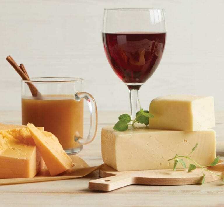 A glass of red wine and a glass mug of mulled cider with blocks of cheddar and other cheese for wine and cheese pairings.