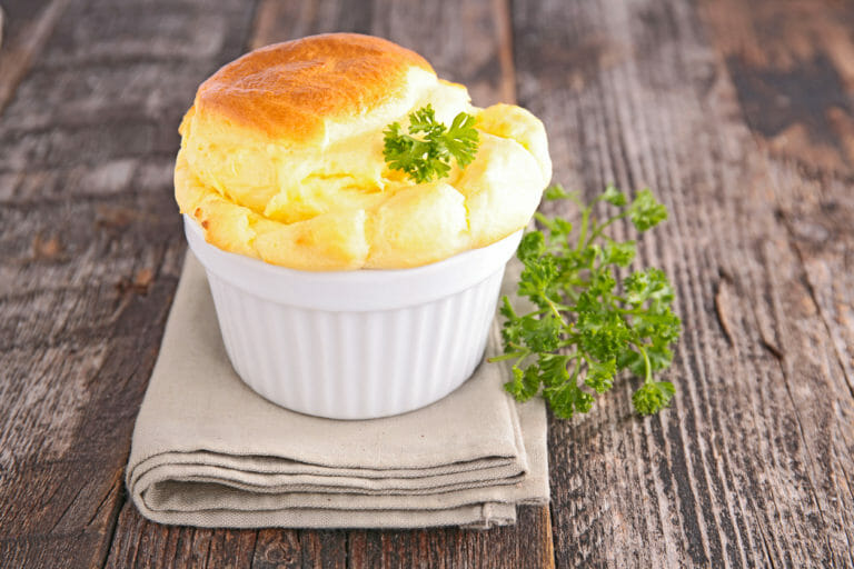 An individual cheese souffle in a white cup with a natural napkin and parsley garnish displayed on a rough barn board table.
