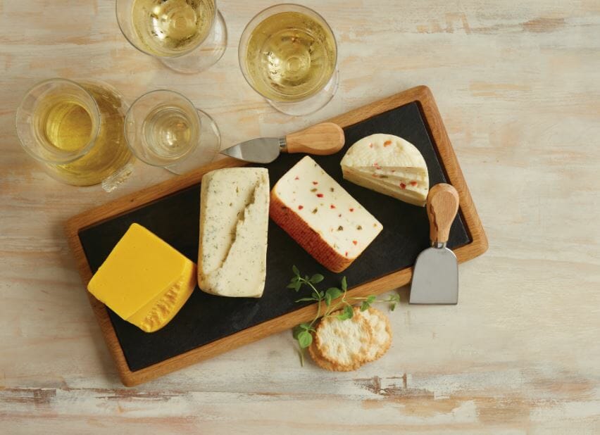 A wooden and slate platter displaying four varieties of cheese, serving utensils, crackers, and glasses of white wine.