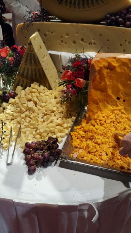 Display table with blocks and sample pieces of 2018 World Championship cheese with tongs, grapes and vased flowers.